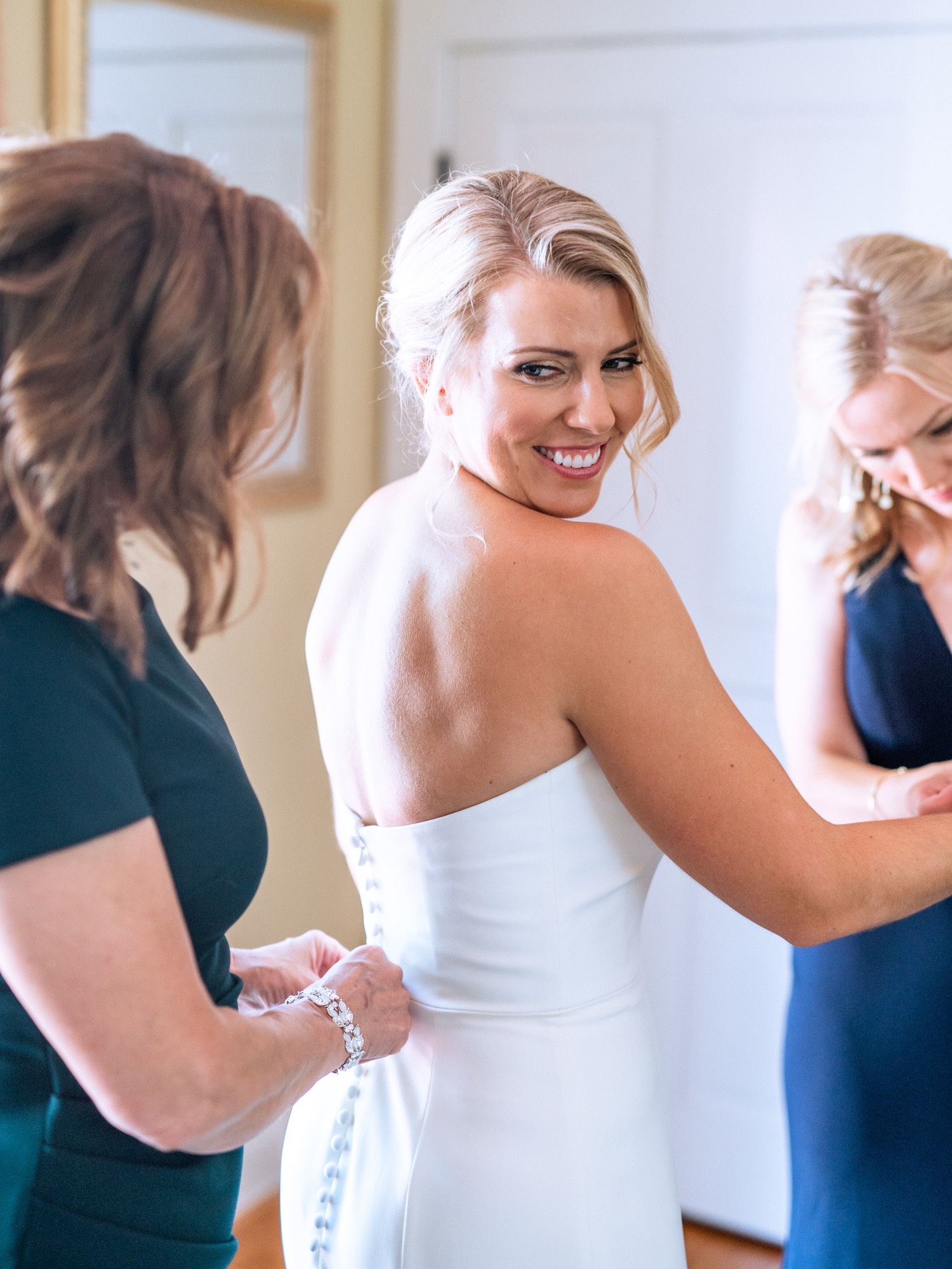 Bride finishes putting her wedding dress on and gets help from mother and maid of honor to button her dress and bracelet before the ceremony in Richmond, Virginia