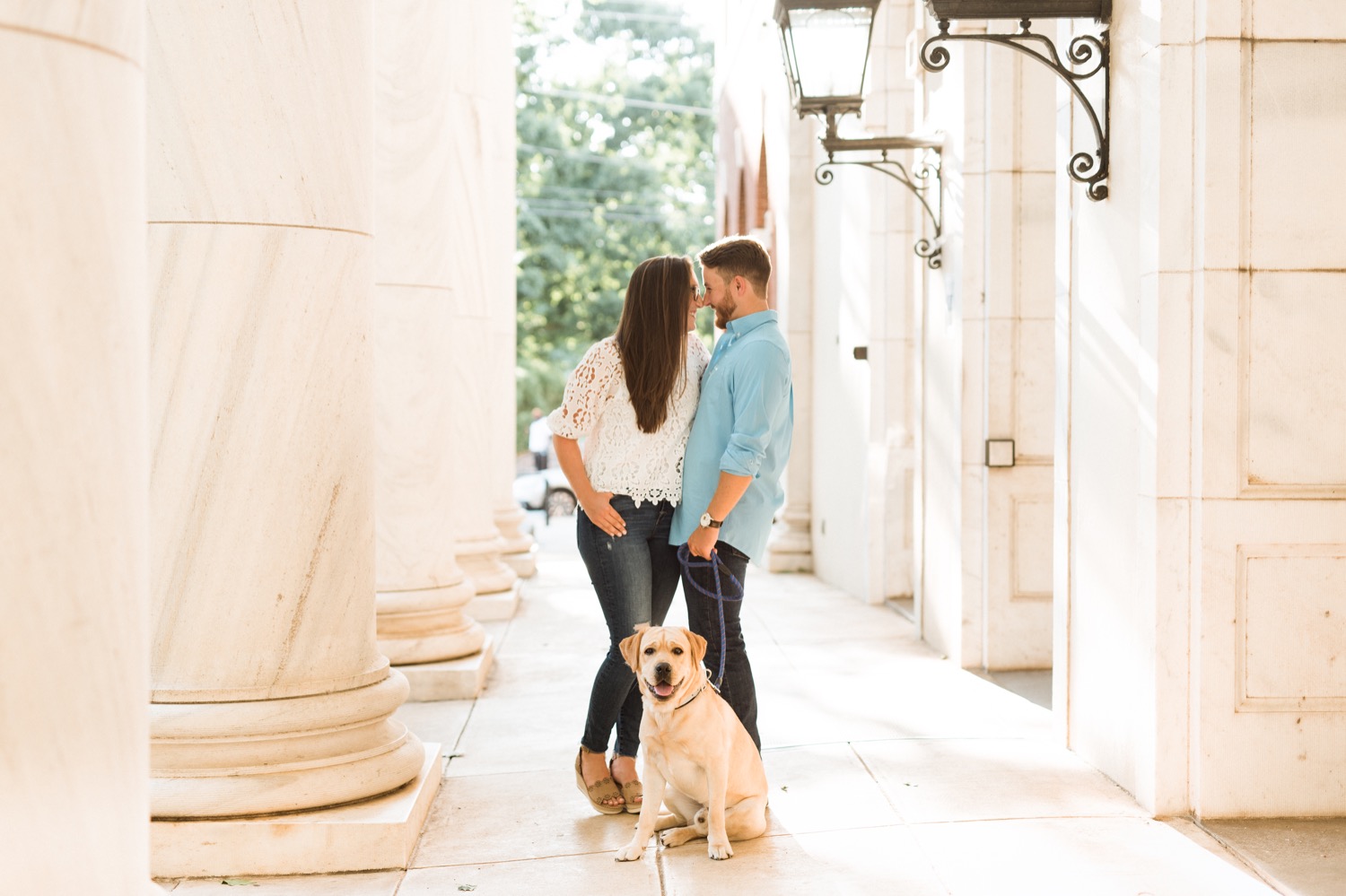 engaged couple kissing while dog sits at their feet and looks right at the camera