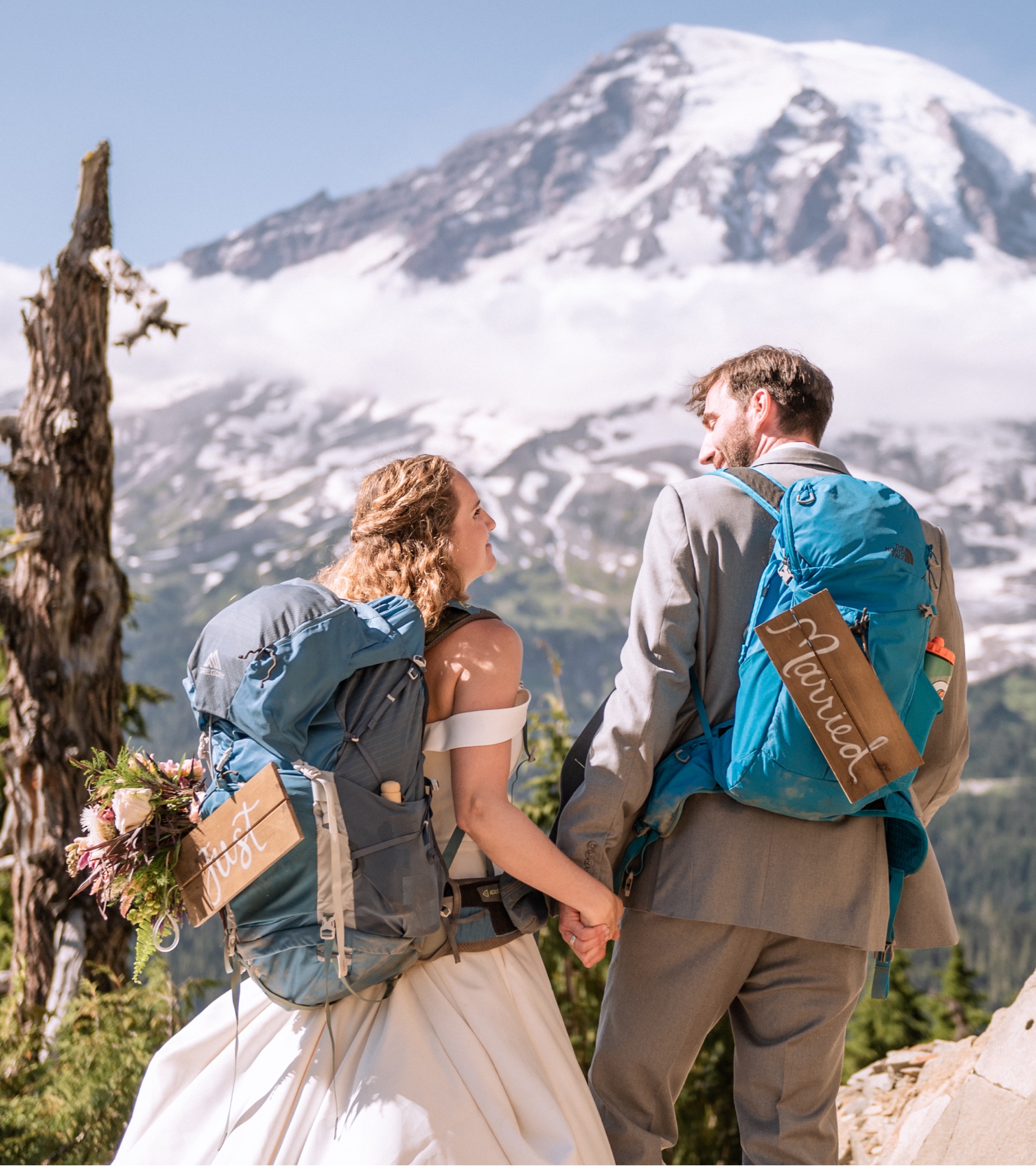 bride and groom in dress and tux with their hiking backpacks on with mt rainier in the background