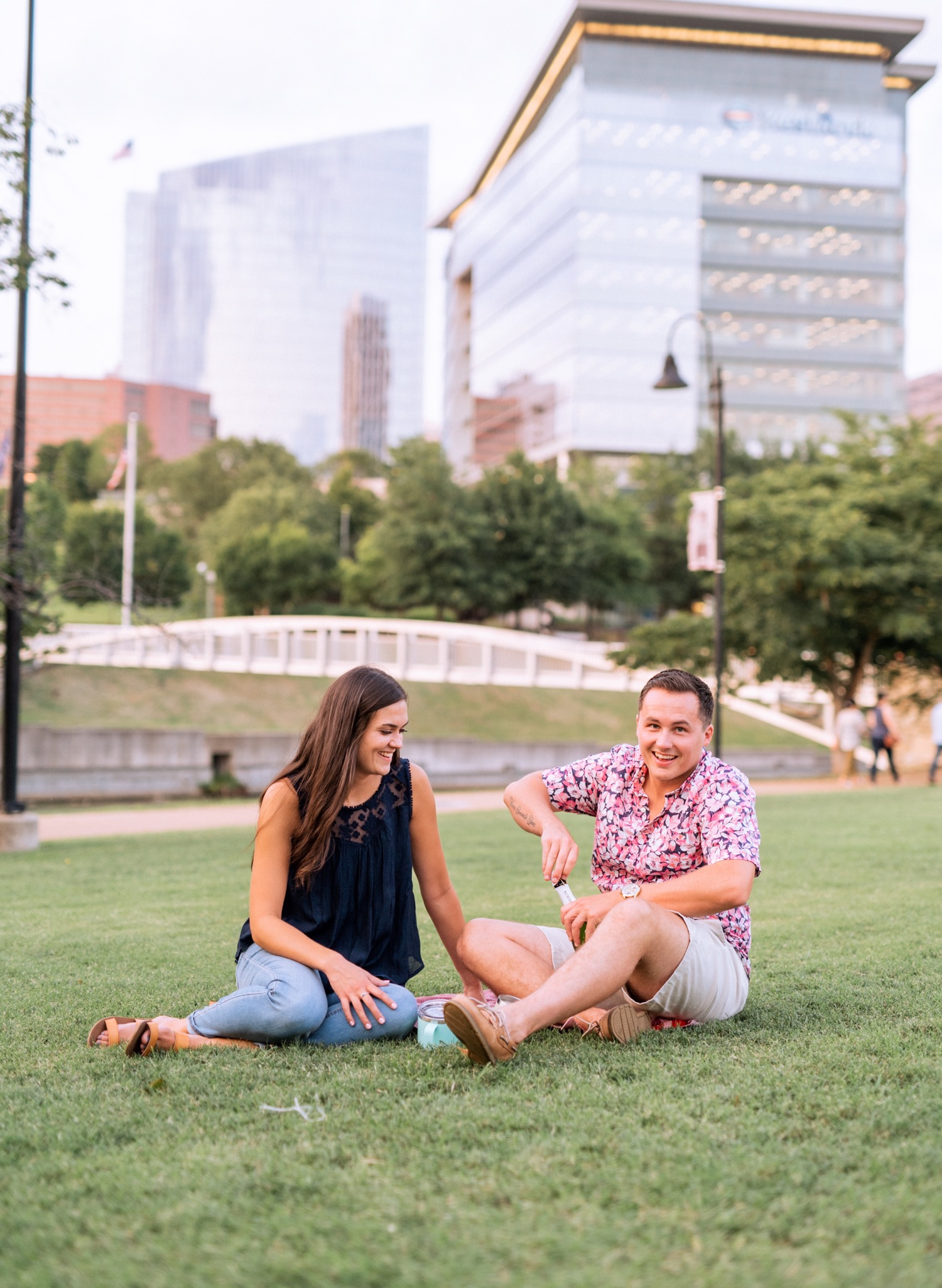 engaged couple sitting and popping champagne bottle on picnic blanket with the skyline and park behind them