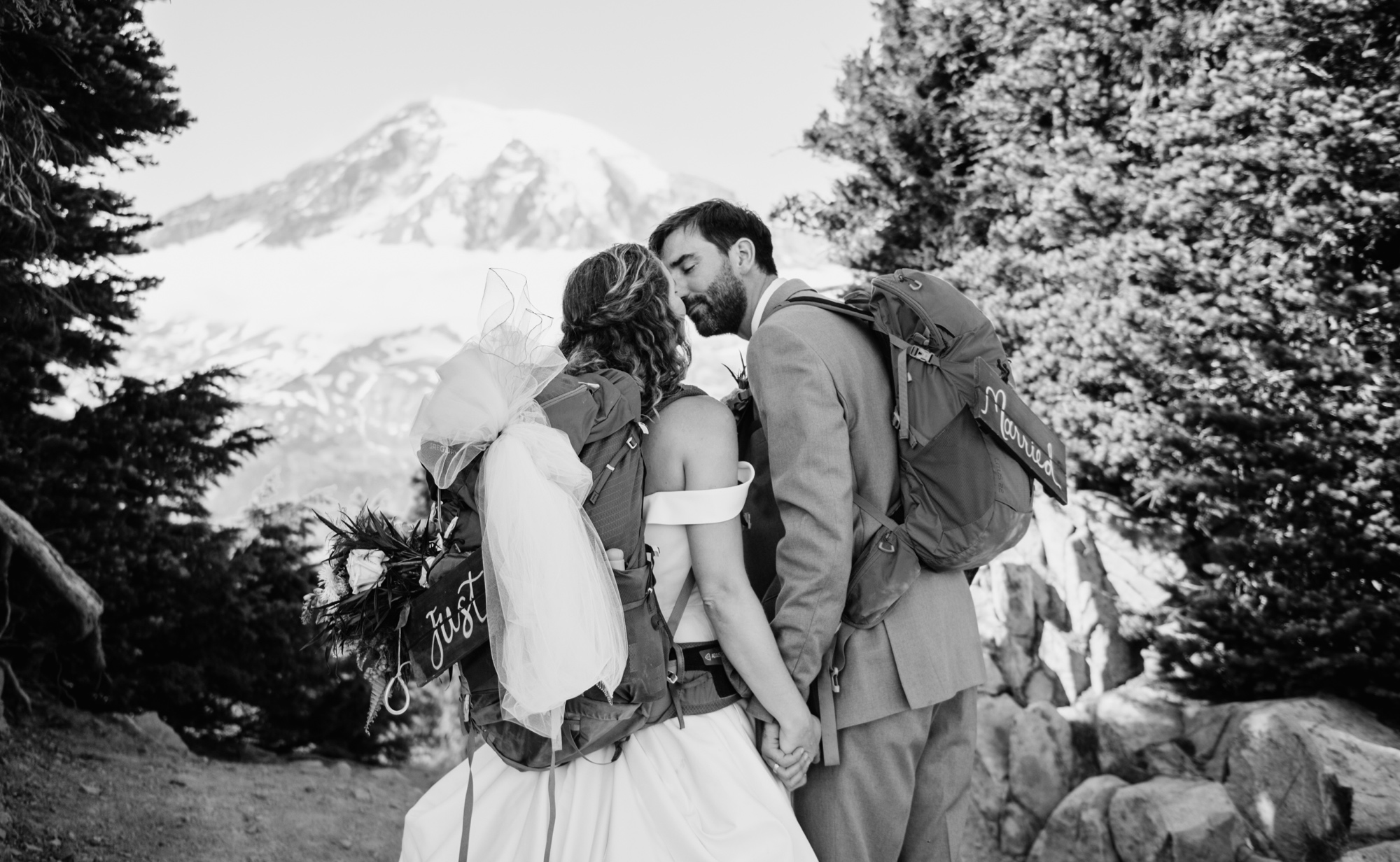 bride and groom kissing in dress and tux with their hiking backpacks on with mt rainier in the background