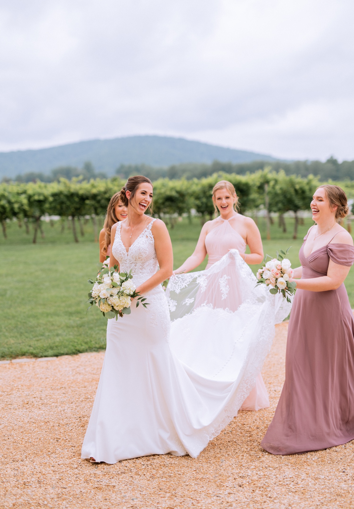 bride and bridal party in mismatching light pink and purple bridesmaids dresses holding white flower arrangements and enjoying themselves