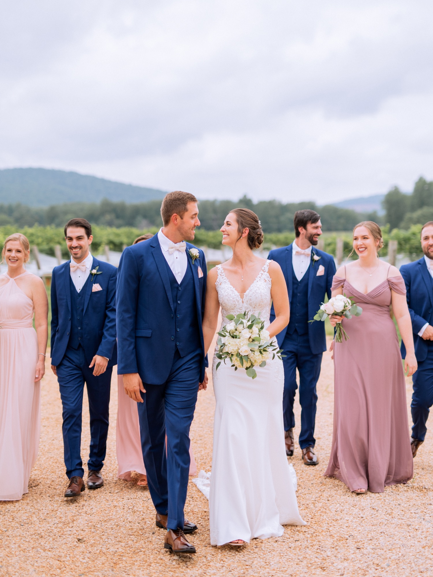 entire wedding party wearing light pink and purple dresses and navy blue tuxes posing and walking with the bride and groom on their wedding day